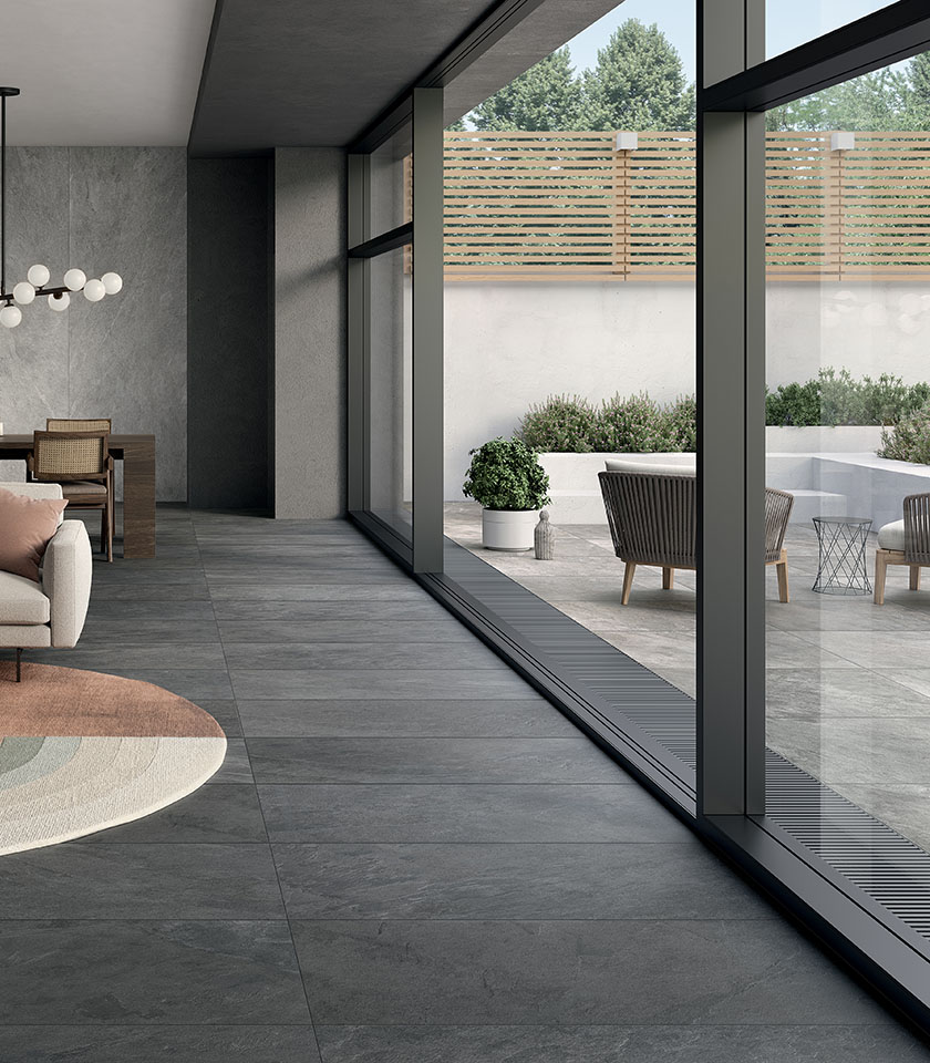 Espace contemporain avec sol effet pierre grise en continuité entre intérieur et extérieur, grandes baies vitrées et terrasse au mobilier épuré.